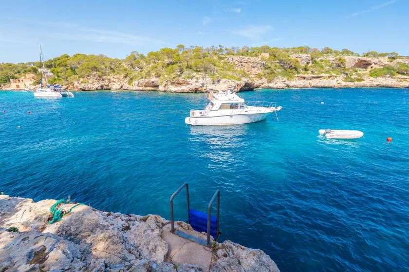 A rocky shoreline overlooks a bright blue bay with boats anchored offshore.
