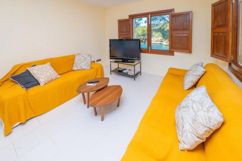 A bright living room with yellow seating, a television, and open wooden shutters.