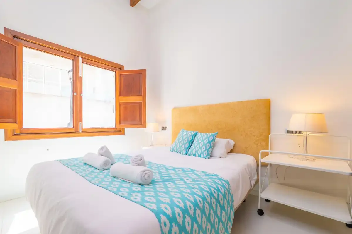 A bedroom with a bed covered in turquoise patterned bedding beside an open window.