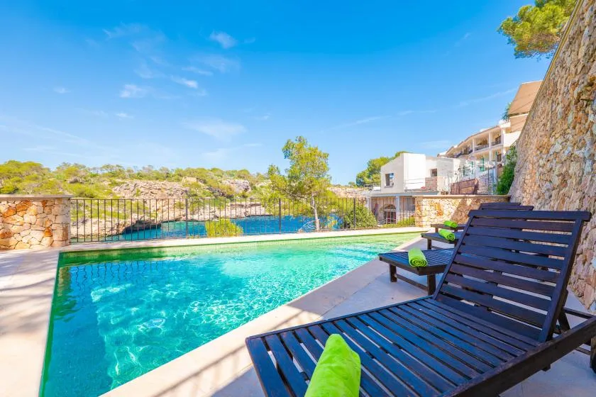 An outdoor swimming pool beside dark lounge seating, with buildings and greenery in the background.