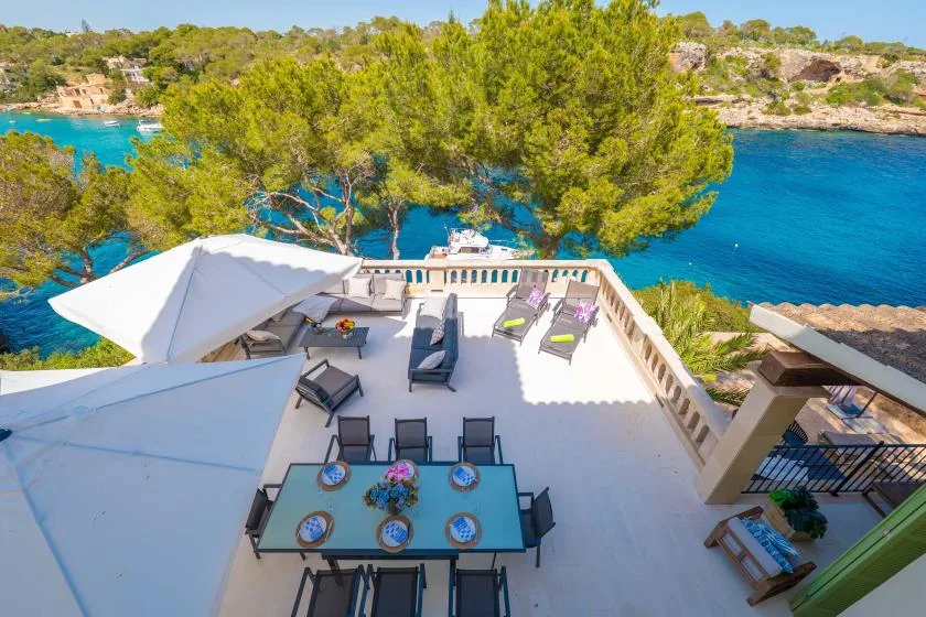 A seaside terrace with outdoor seating overlooks a bright blue bay.