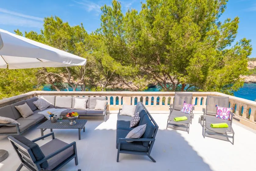 A bright outdoor terrace has cushioned seating, a low table, and leafy trees.