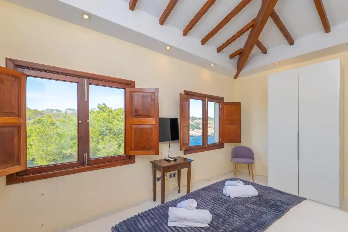 A bright bedroom with wooden ceiling beams and windows overlooking greenery.