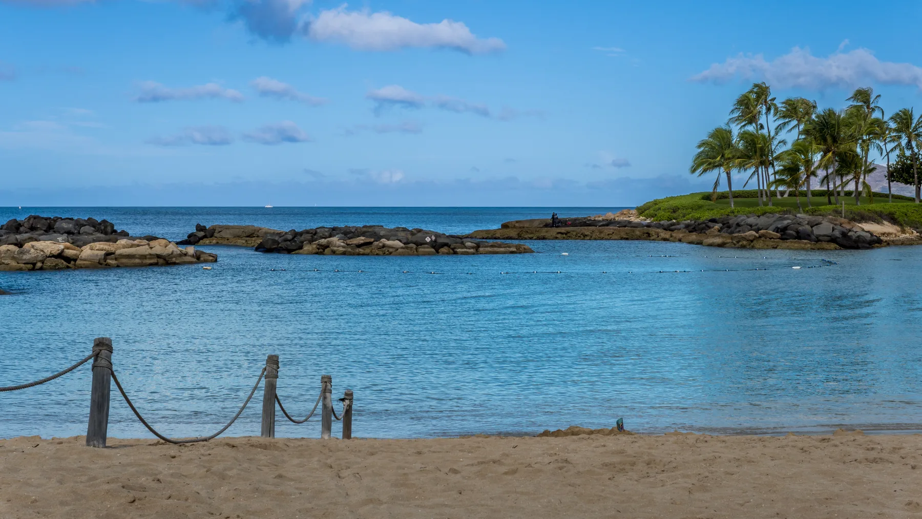 Una playa de arena da a aguas azules tranquilas y una orilla rocosa bordeada de palmeras.