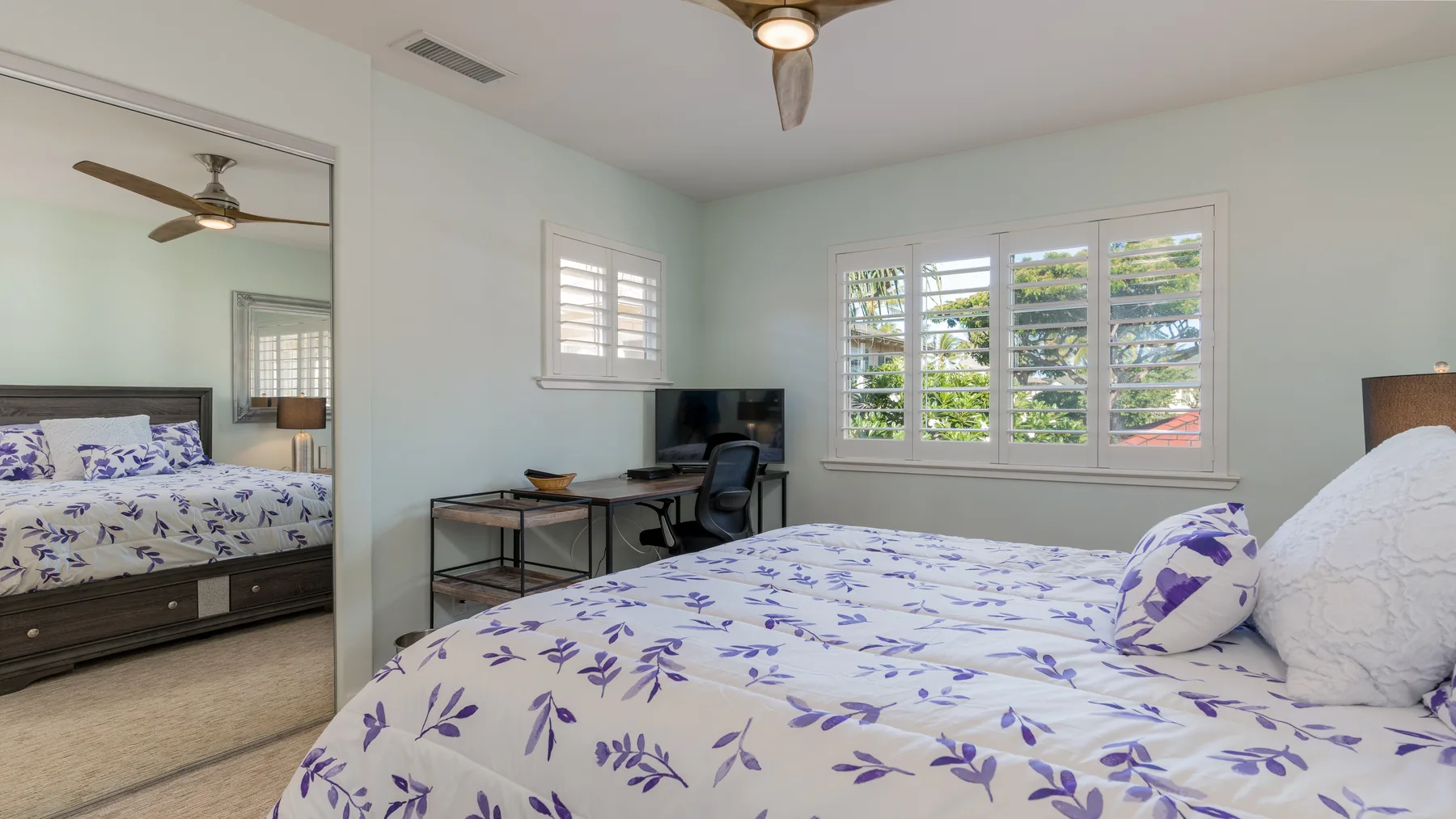 Un dormitorio con una cama de ropa de cama floral morada y un escritorio junto a las ventanas.