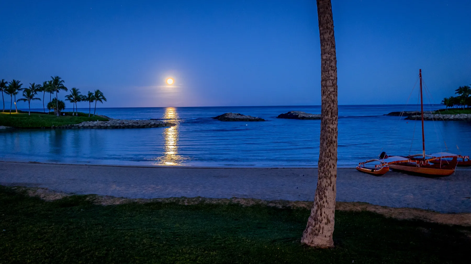 Una playa al anochecer da al mar en calma, con la luna reflejada en el agua.