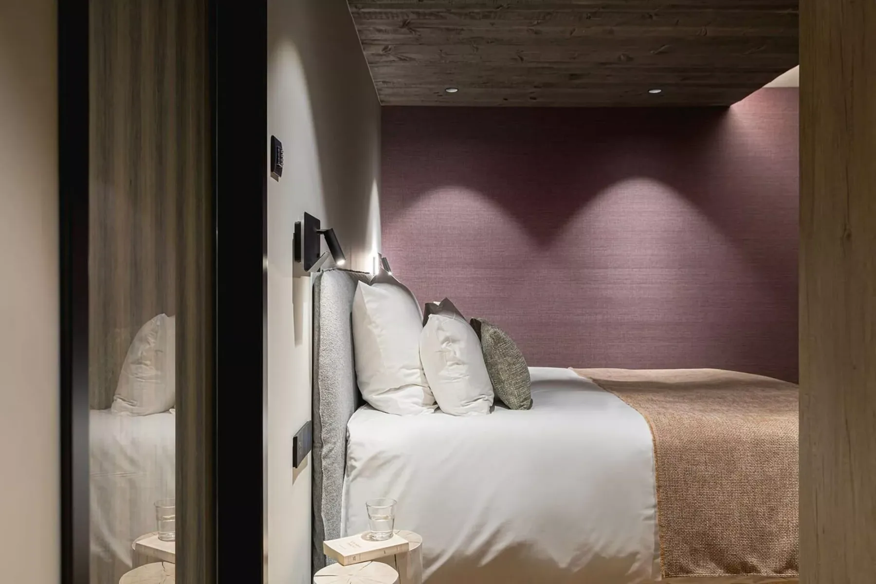 A bedroom with a neatly made bed against a softly lit mauve wall.