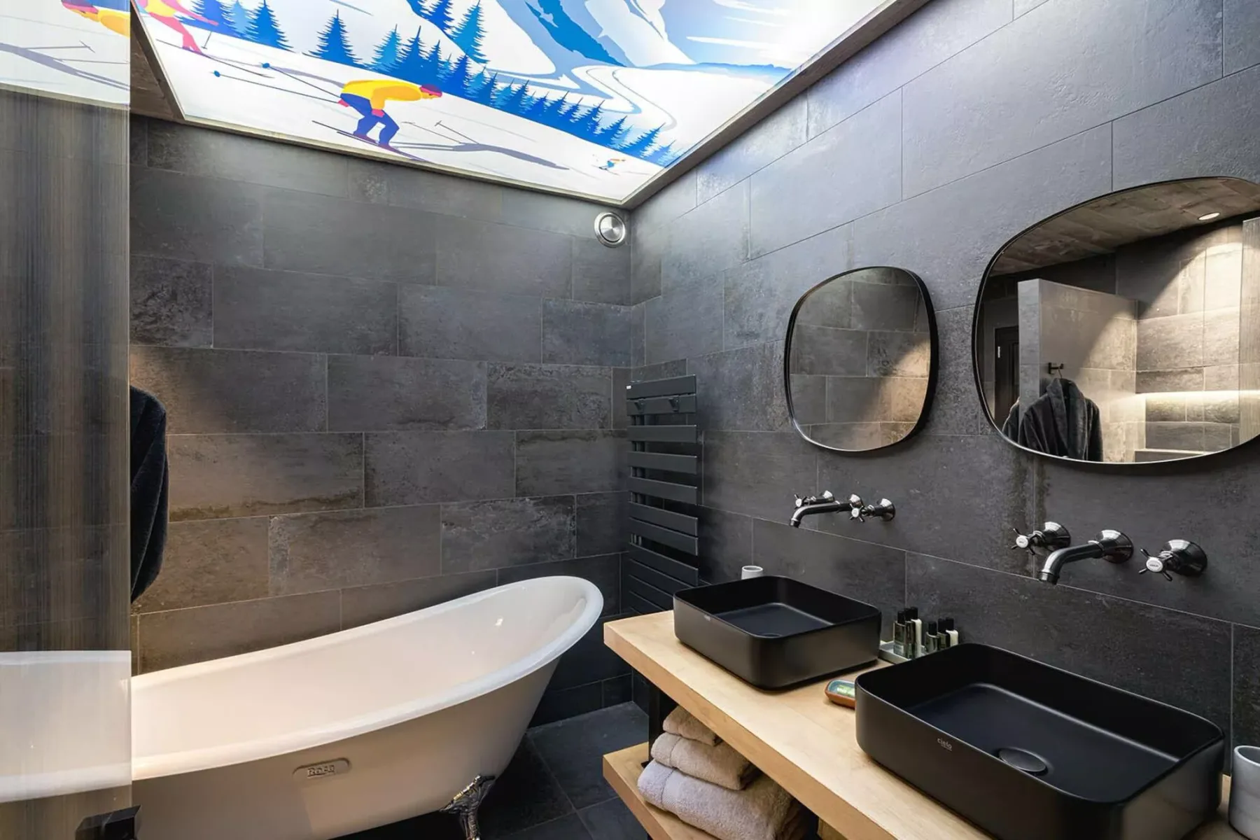 A dark-tiled bathroom with a bathtub, two sinks, and mirrors.