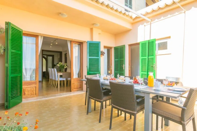 Comedor en un patio cubierto con mesa y sillas junto a persianas verdes abiertas.