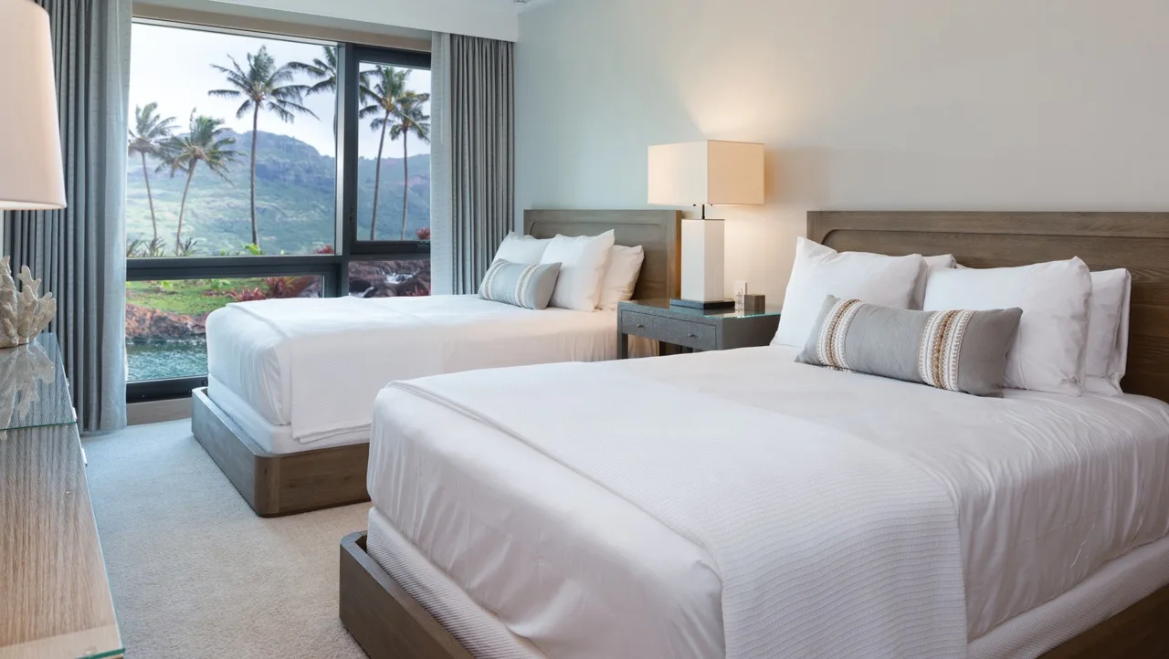 A bedroom with two neatly made beds beside a large window overlooking palm trees.
