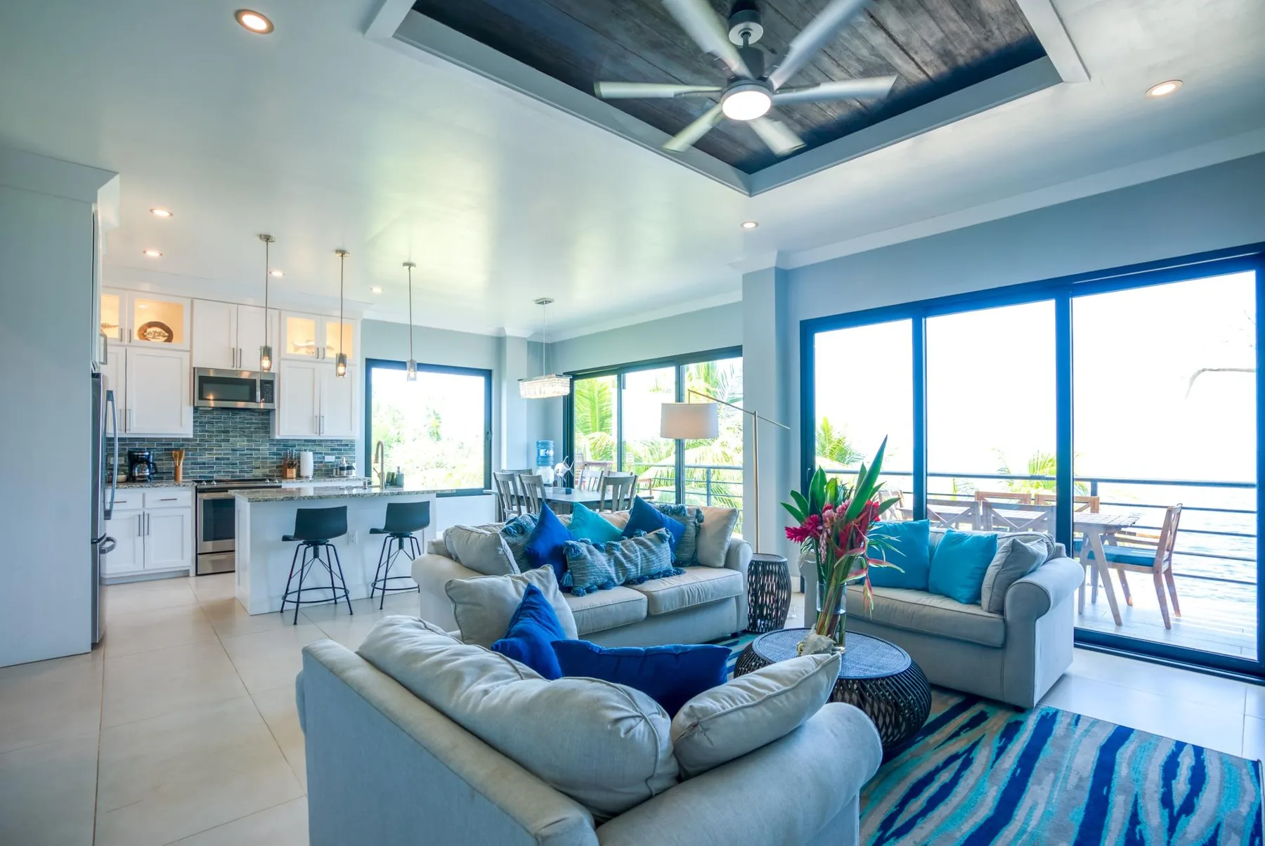 A bright open-plan living room has seating beside large windows and a kitchen.