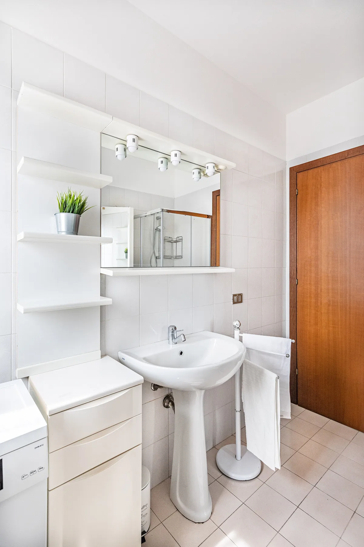Un baño luminoso con lavabo de pedestal, espejo y azulejos blancos.
