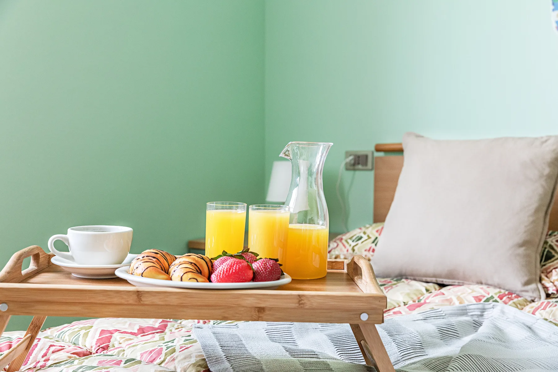 Una cama con una bandeja de desayuno con bollería, fruta, zumo y una taza.