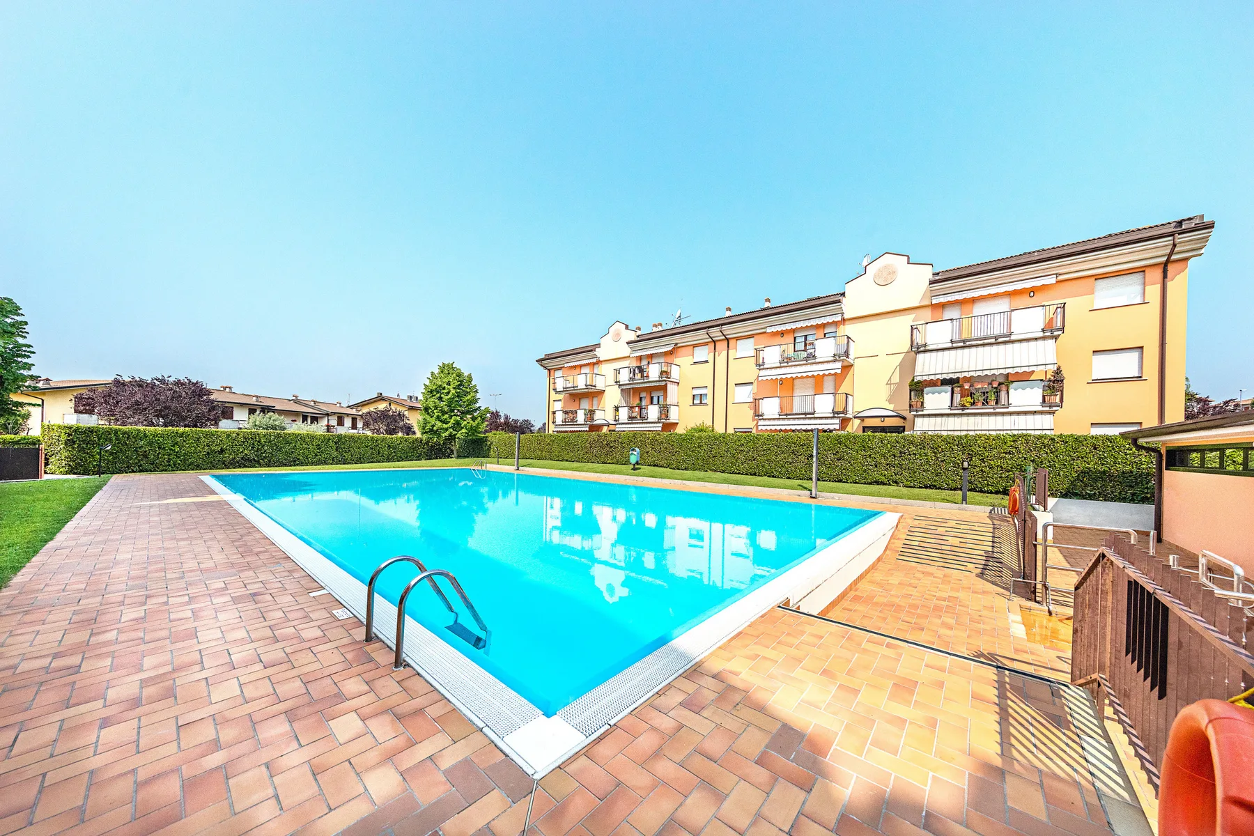 Una piscina al aire libre junto a una terraza pavimentada y un edificio de varias viviendas.