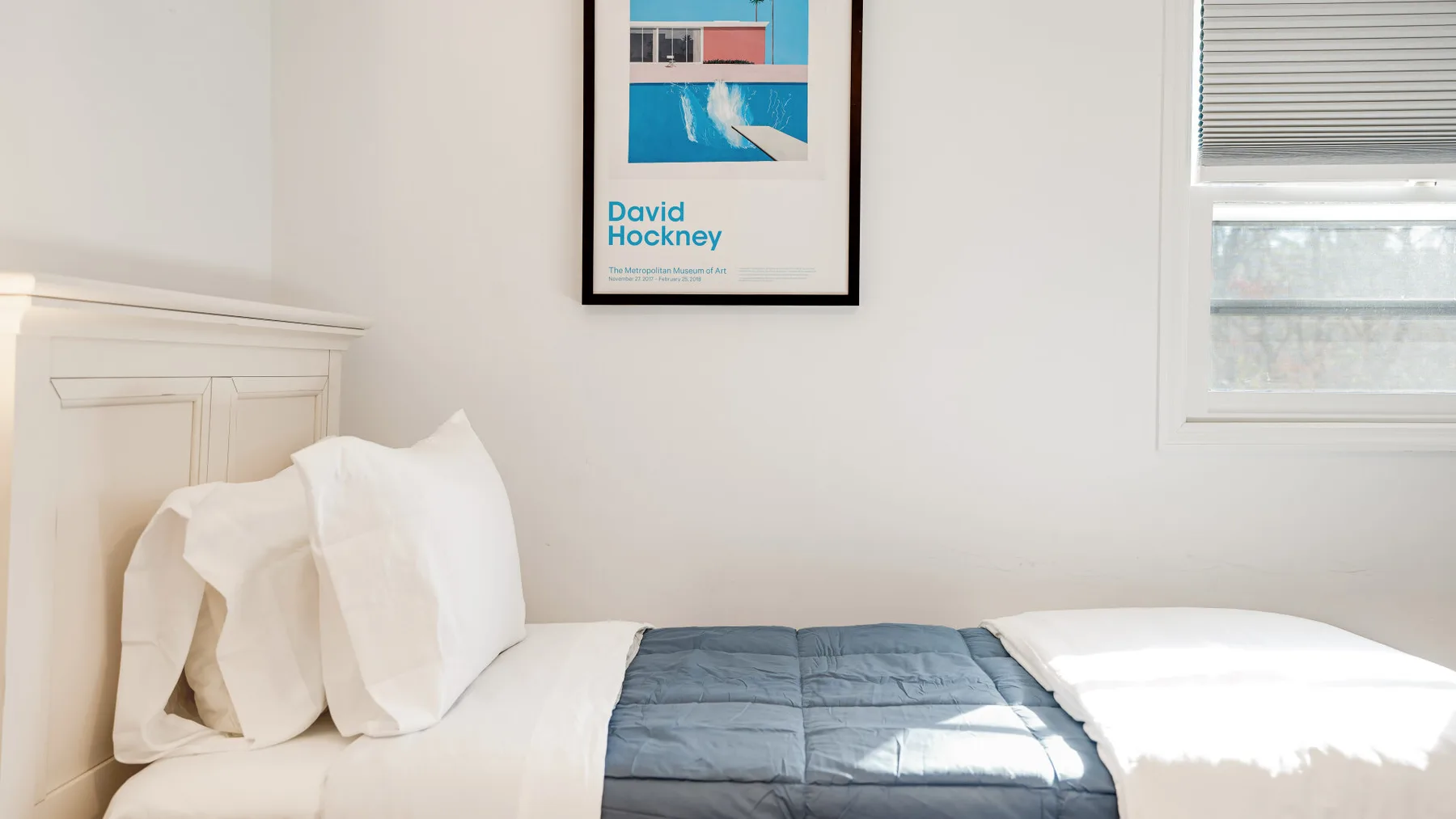 A bright bedroom with a bed beneath a framed art print.