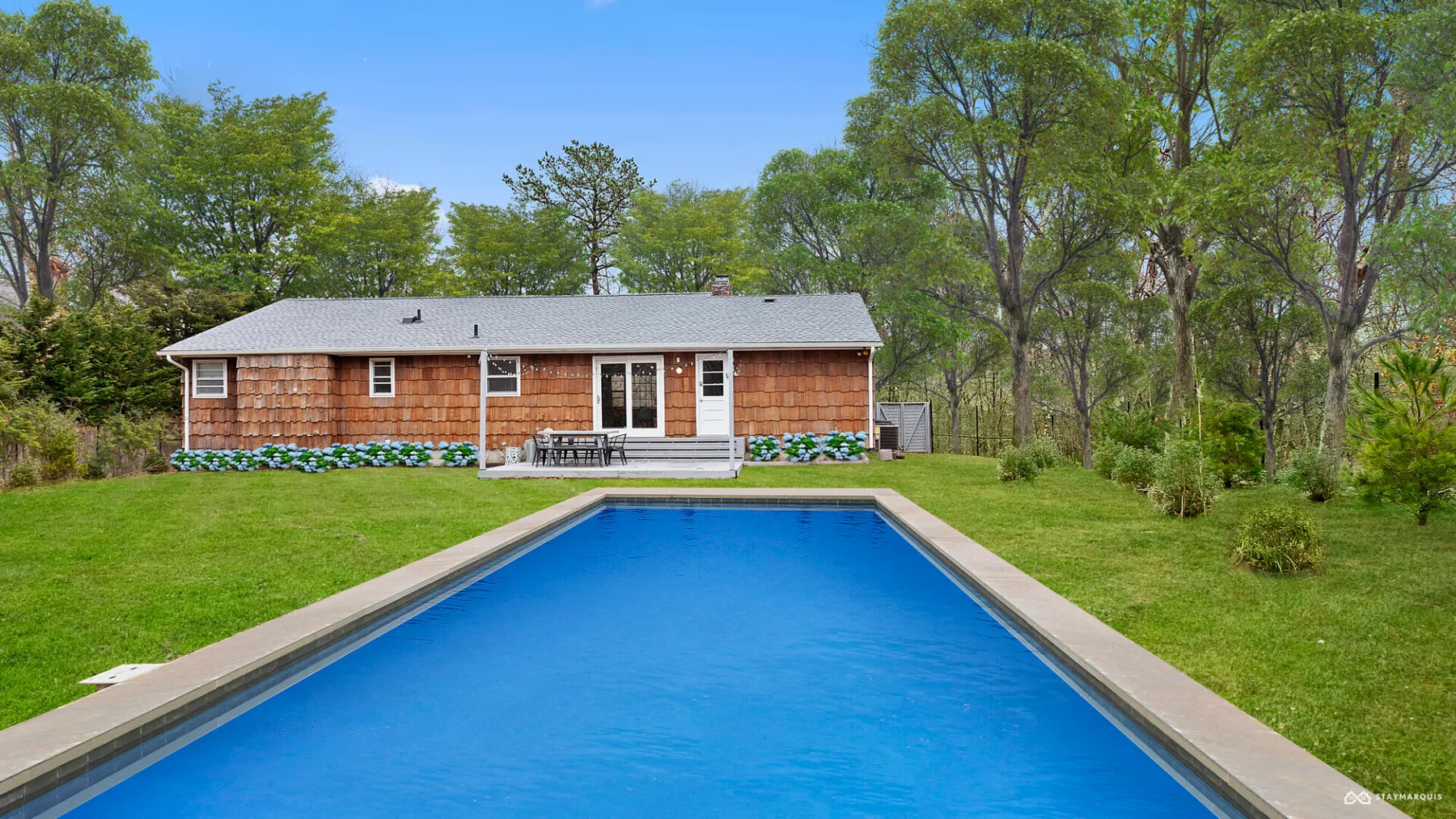 A swimming pool stretches across the lawn toward a single-story house surrounded by trees.