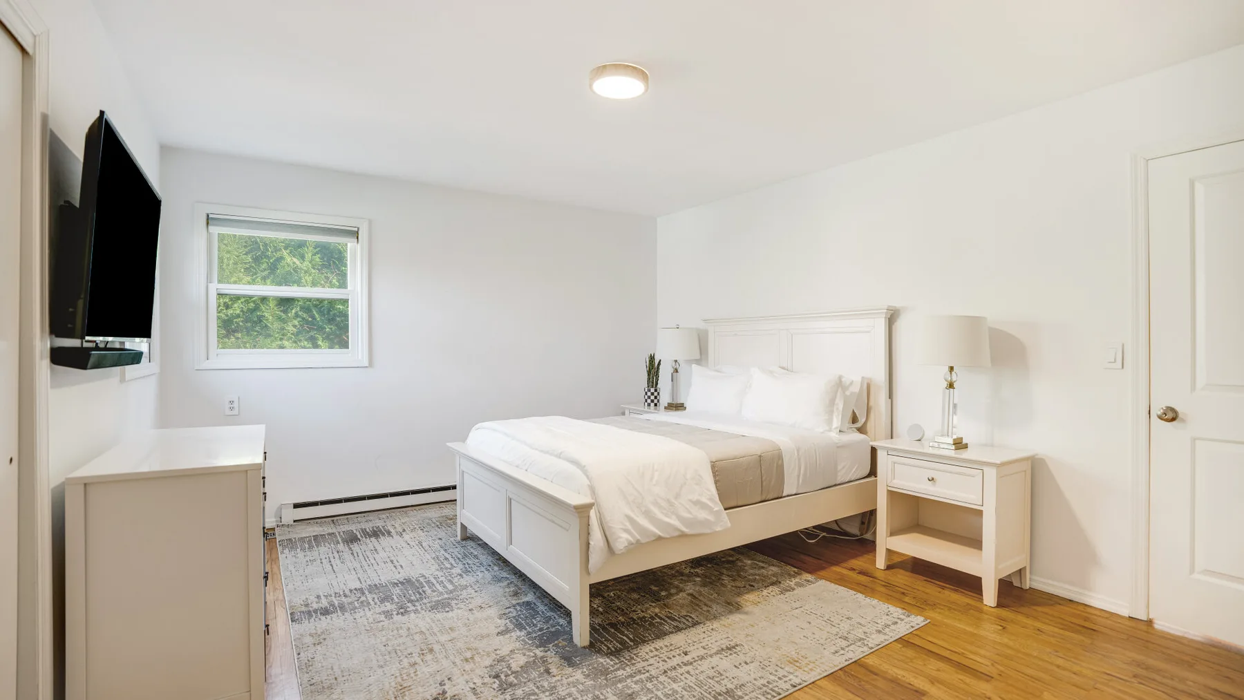 A bright bedroom with a white bed and a window overlooking greenery.