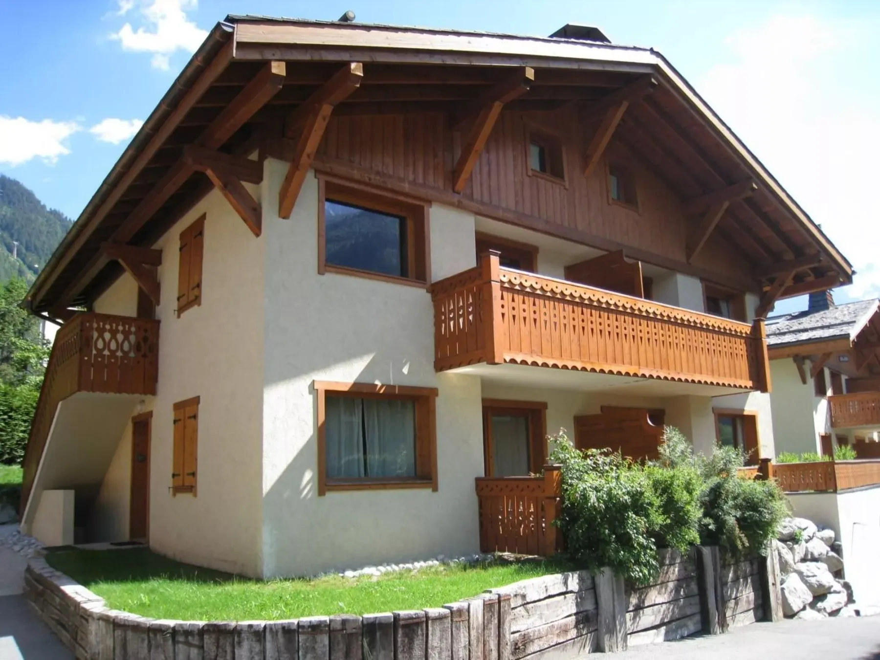 A chalet-style house with wooden balconies stands beside a low stone wall.