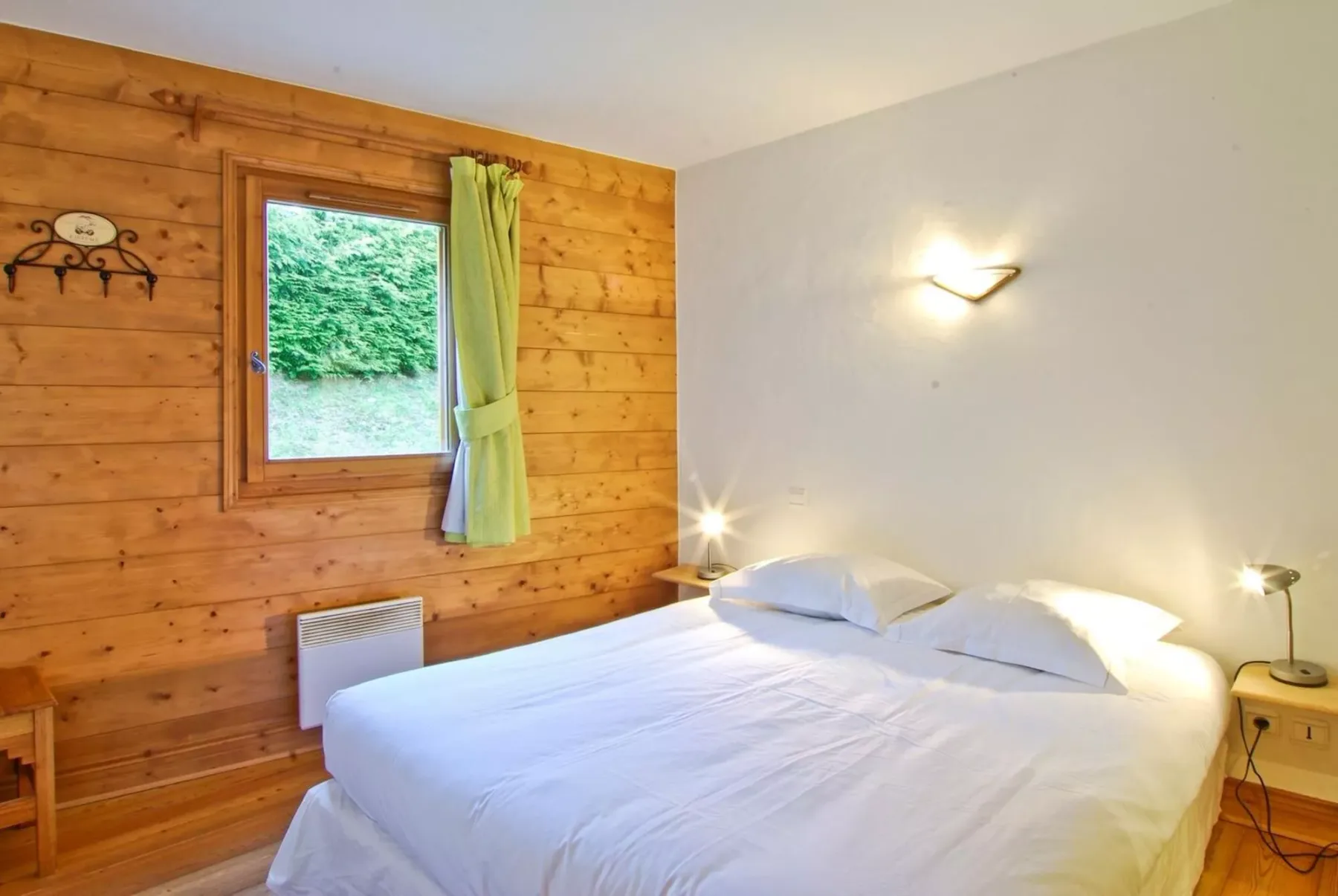 A bedroom with a white bed, wooden walls, and a window overlooking greenery.