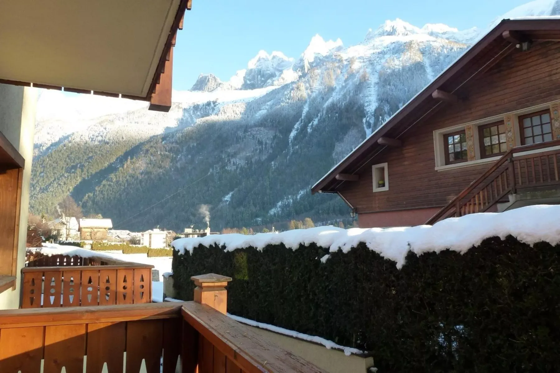 A wooden balcony overlooks snow-covered mountain peaks beyond nearby chalet-style buildings.