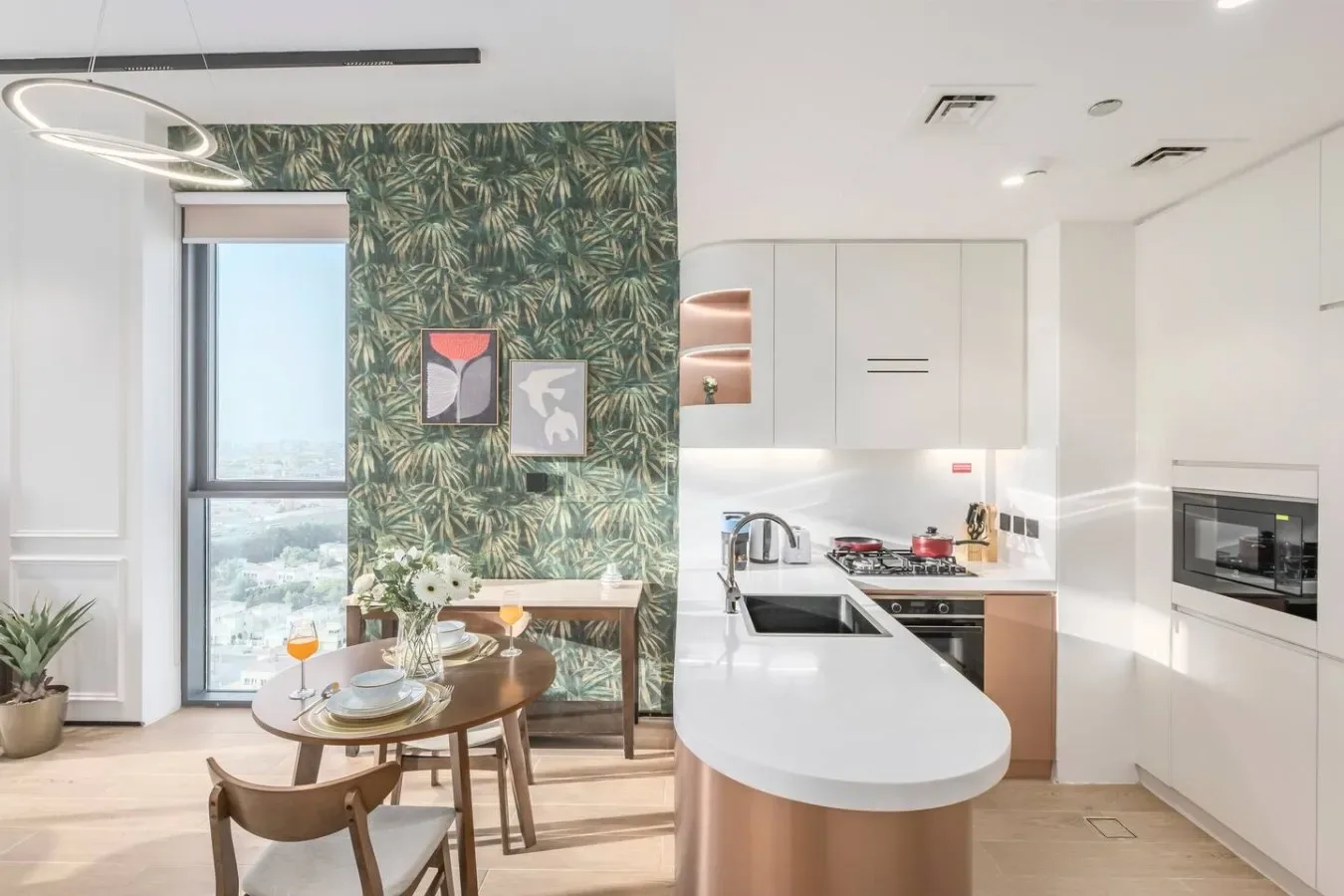 Una cocina moderna y zona de comedor con armarios blancos y una mesa junto a una pared decorativa de hojas.