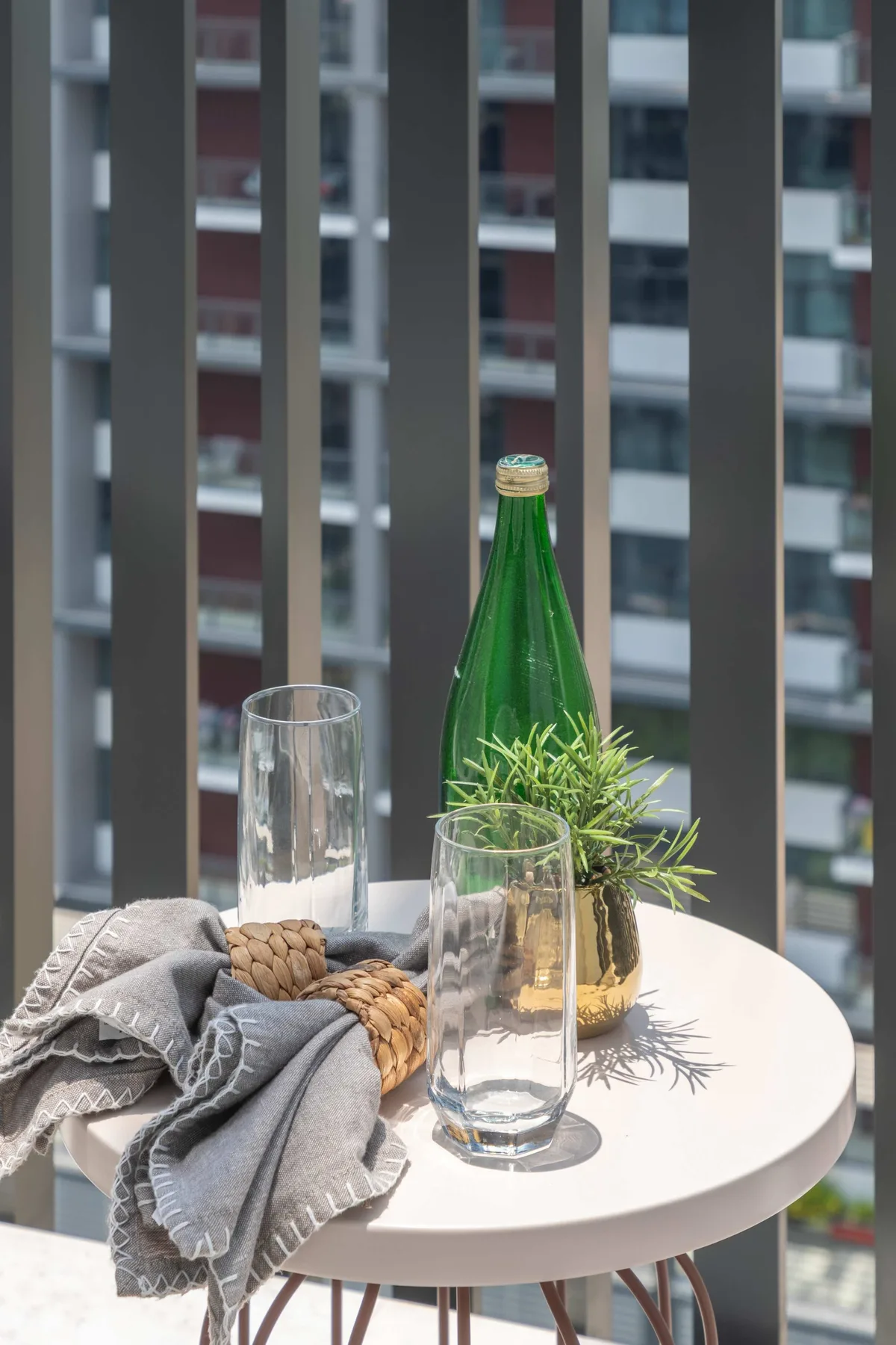 Una pequeña mesa redonda junto a una ventana tiene vasos, una botella verde y una planta.