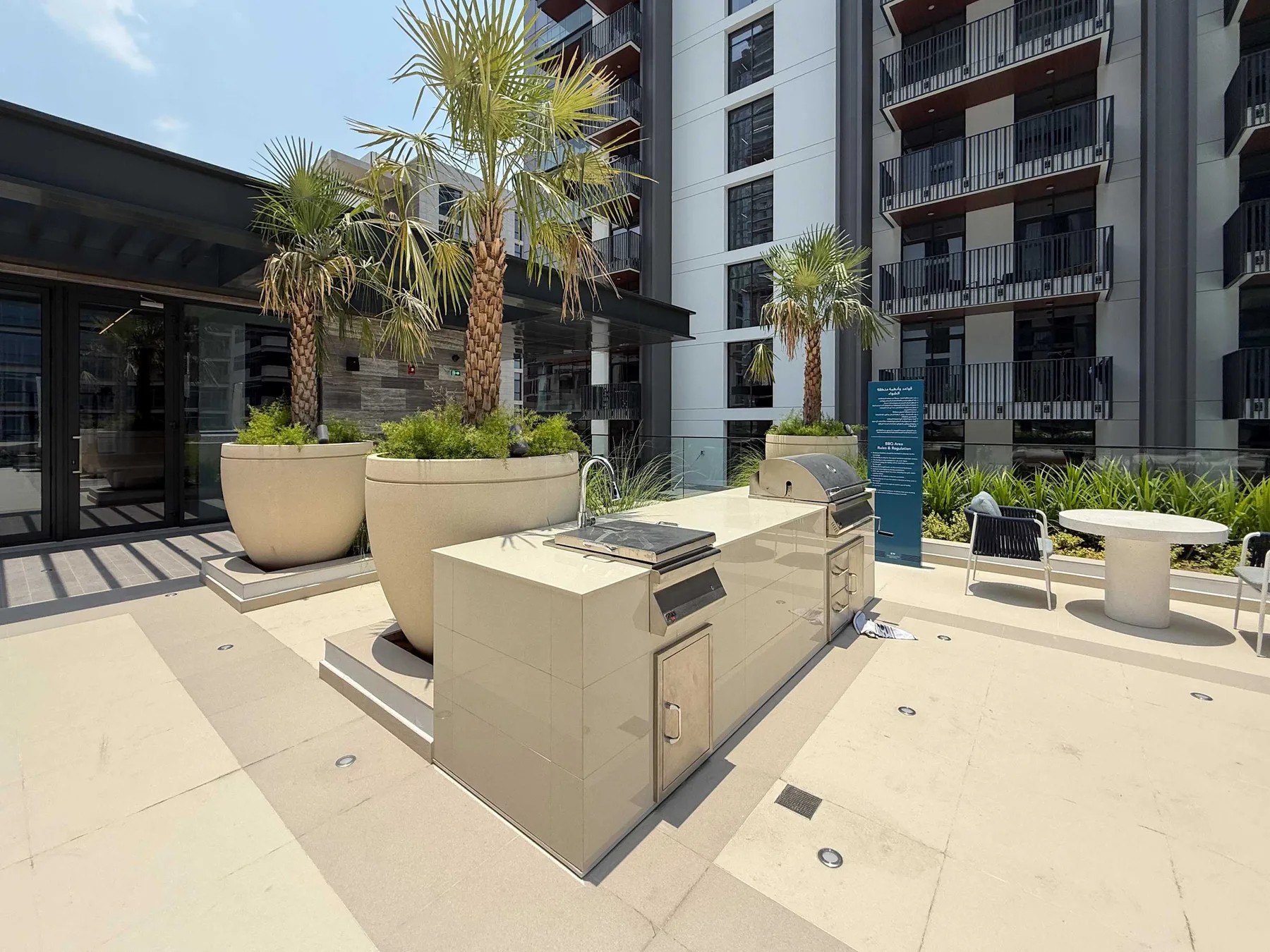 Un patio soleado de apartamentos con palmeras, asientos al aire libre y una parrilla incorporada.