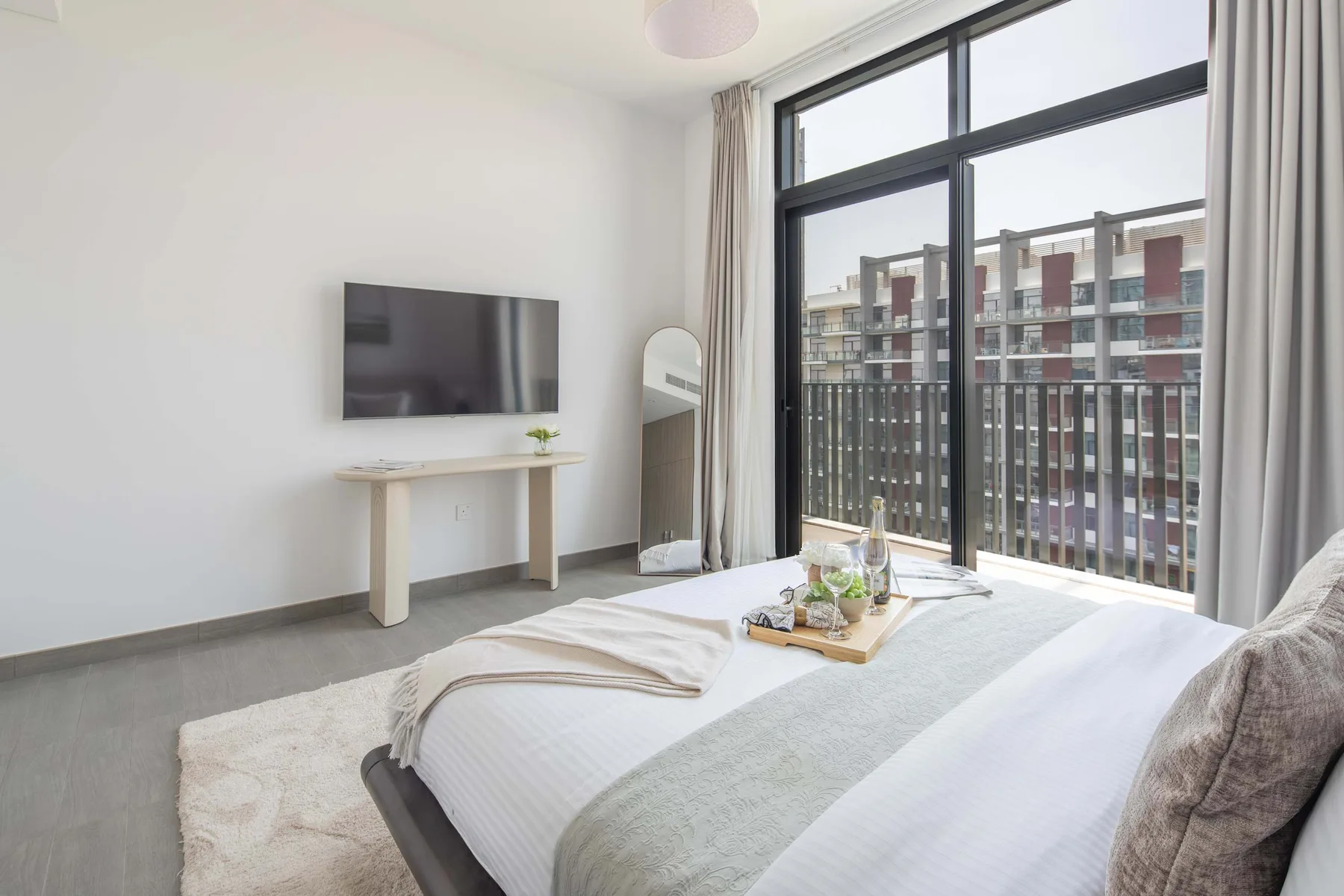 Un dormitorio luminoso con cama, televisor en la pared y ventanas amplias.