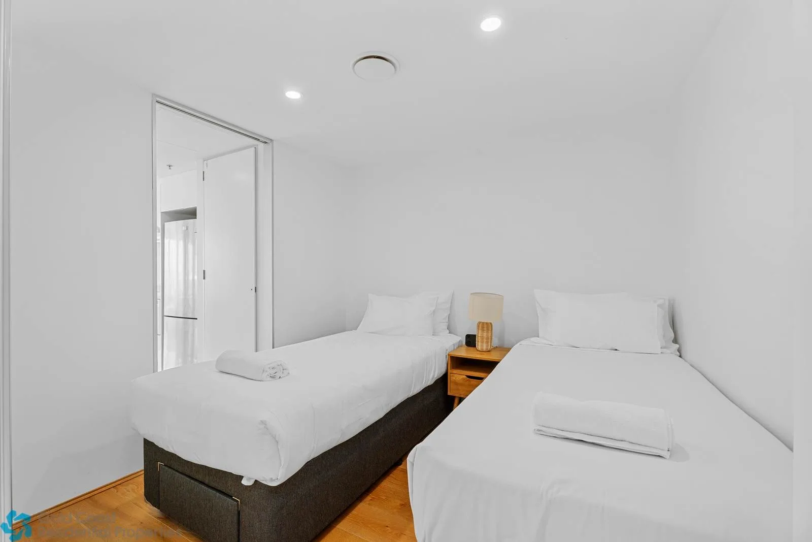 A bright bedroom with two beds, white bedding, and a small bedside table.