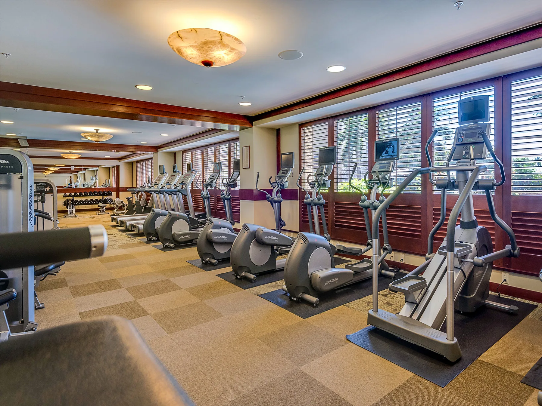 Un gimnasio con máquinas cardiovasculares junto a grandes ventanas.