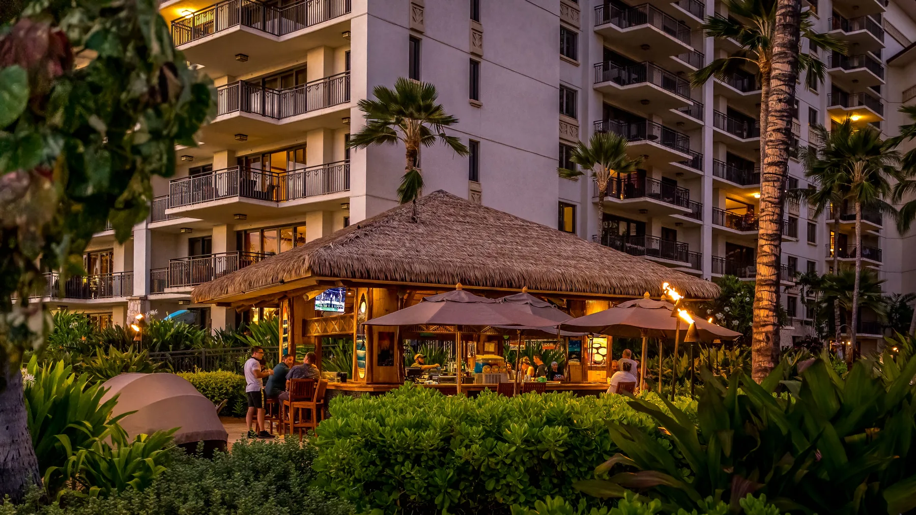 Un restaurante al aire libre con techo de paja está entre plantas tropicales junto a un edificio alto.