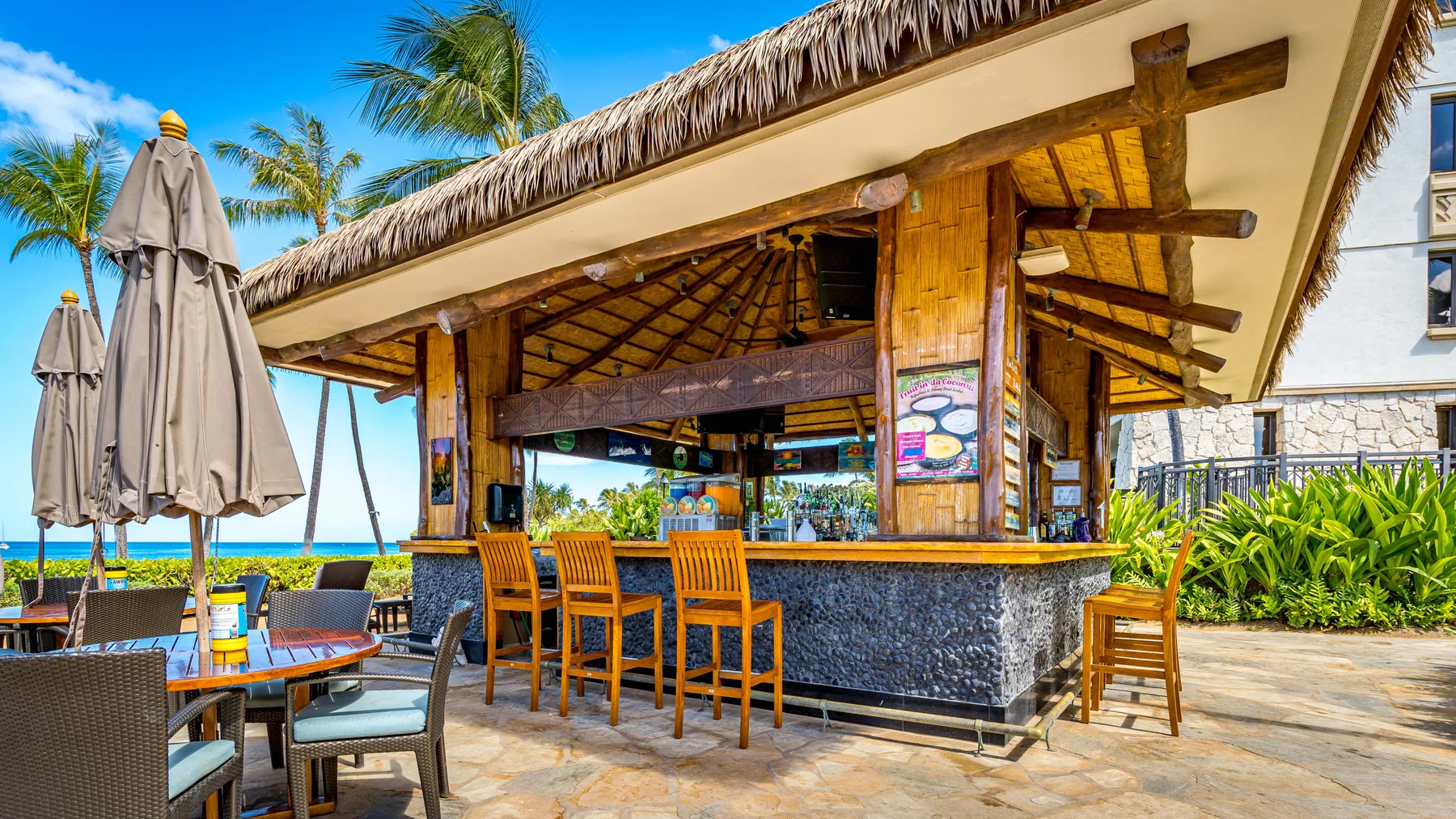 Un bar al aire libre con taburetes y mesas de madera bajo un techo de paja.