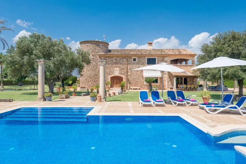 Una piscina está frente a una casa de piedra, con tumbonas y sombrillas cerca.