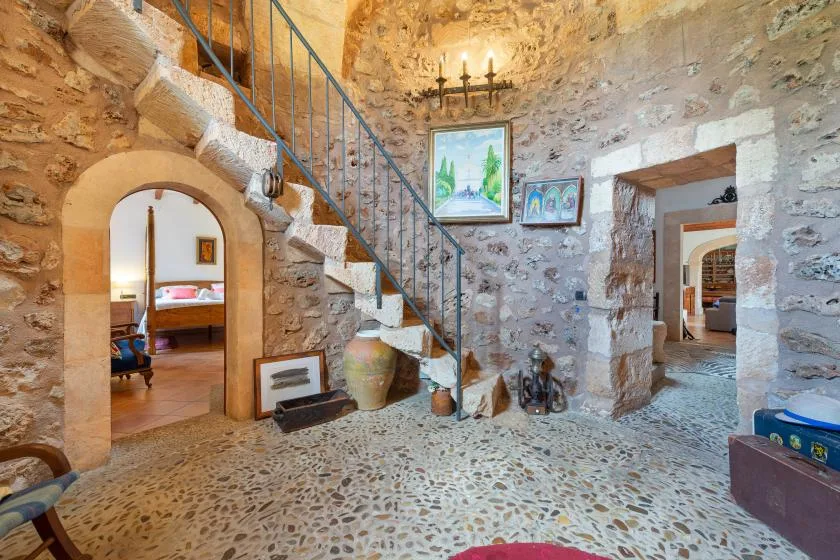 Interior de piedra con una escalera, cuadros enmarcados y puertas arqueadas.