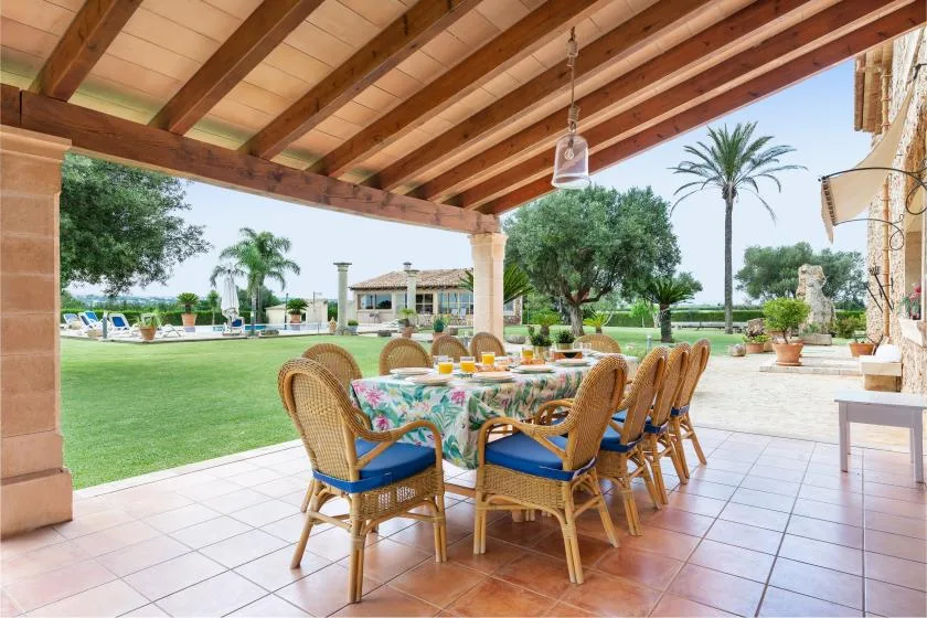 Una zona de comedor exterior cubierta con una mesa larga y asientos de mimbre da a un jardín cuidado.