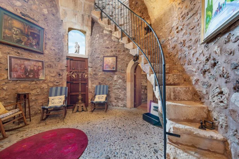 Interior de piedra con una escalera curva, cuadros enmarcados y asientos.