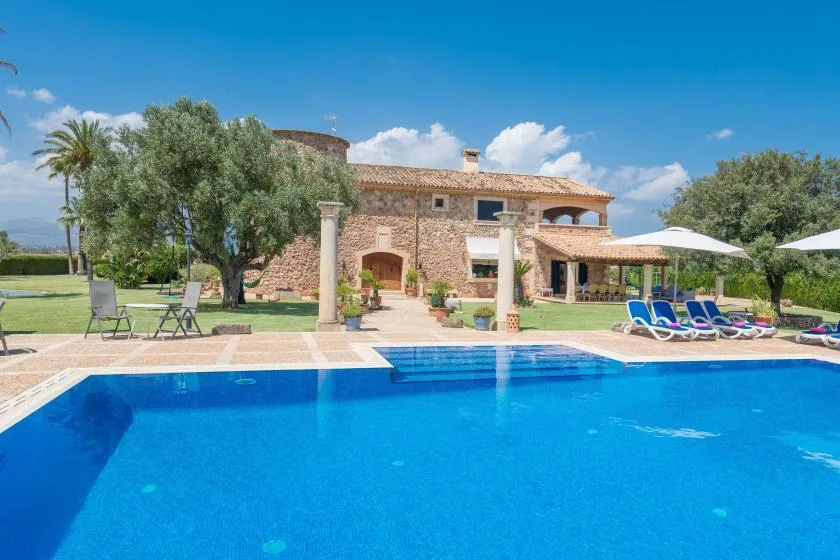 Una piscina está frente a una casa de piedra, con asientos y sombrillas cerca.