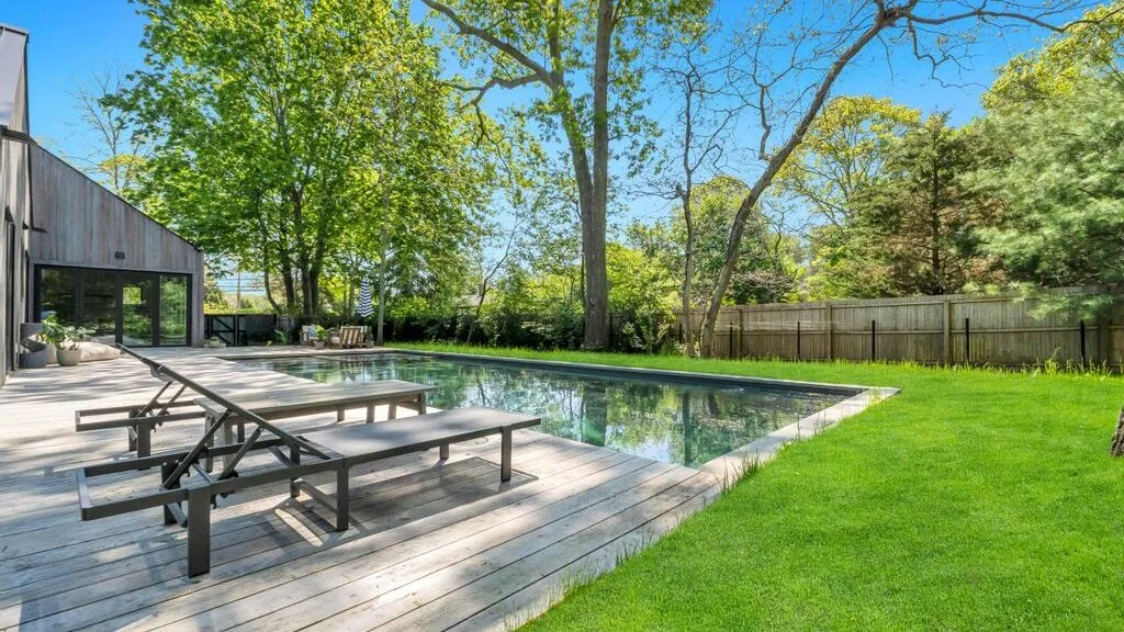 Una piscina en el patio junto a una terraza de madera, césped y árboles.