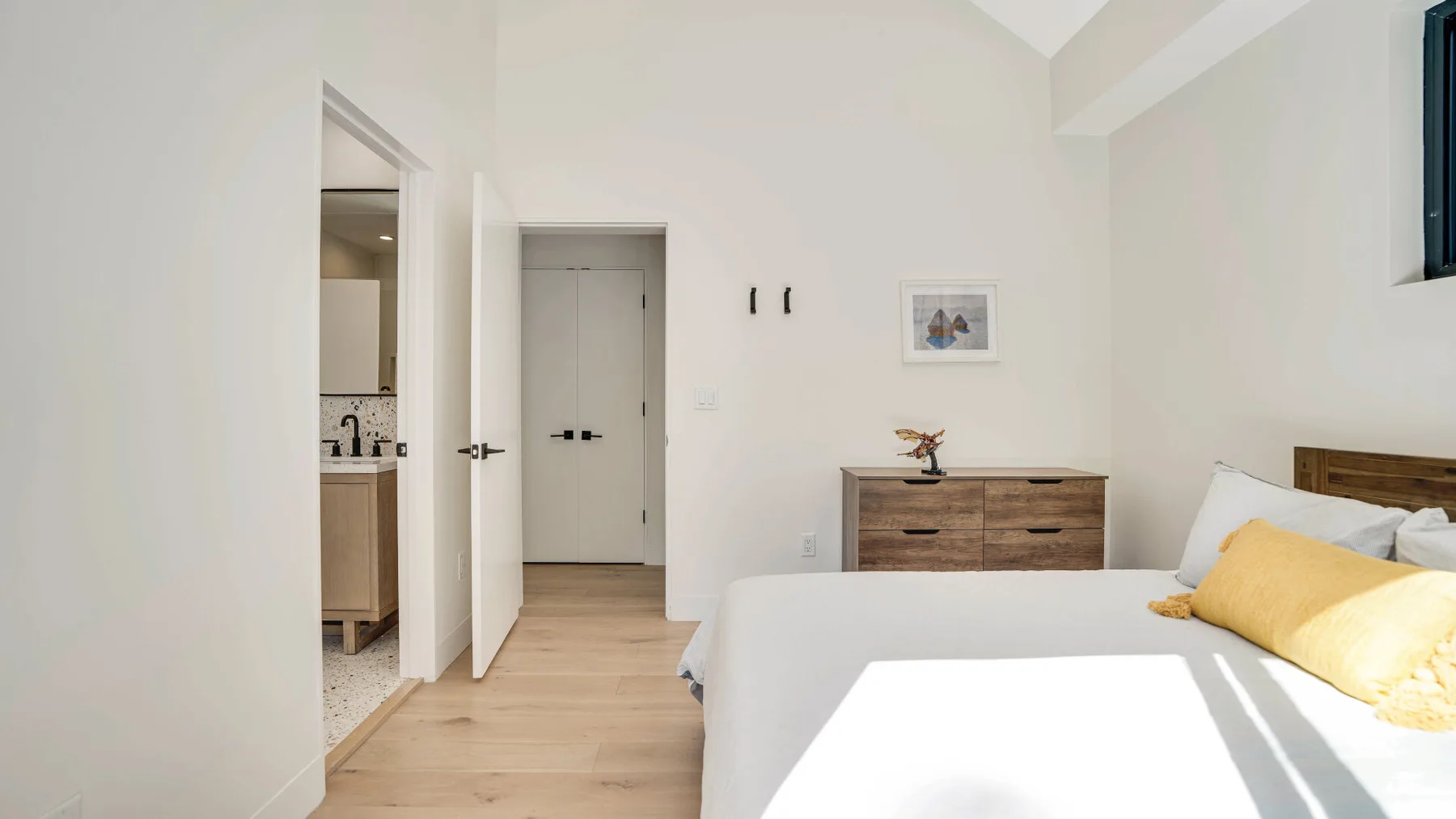 Dormitorio luminoso con cama, cómoda de madera y puerta abierta.