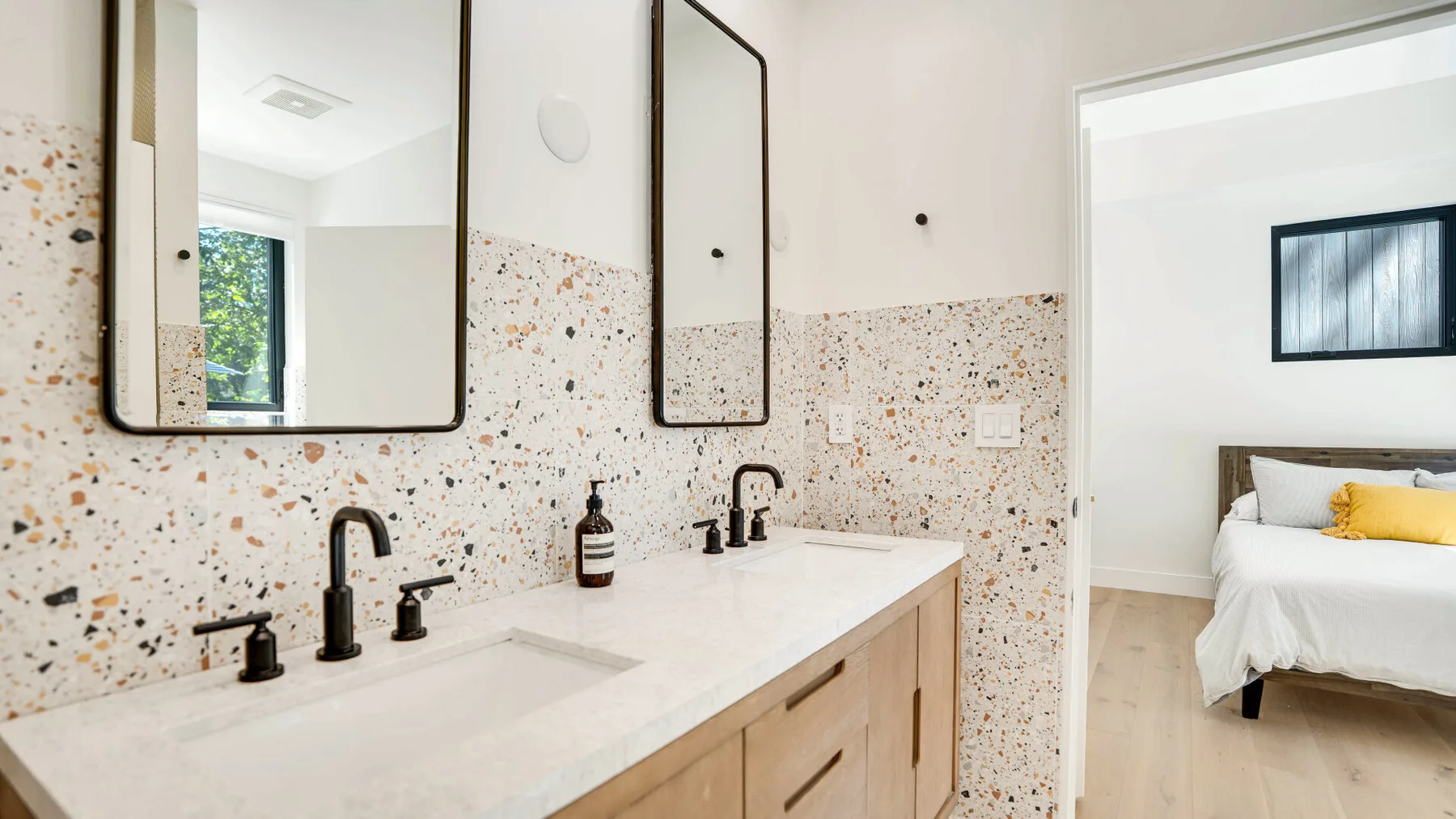 Un baño luminoso con un tocador largo y espejos, con vistas a un dormitorio.