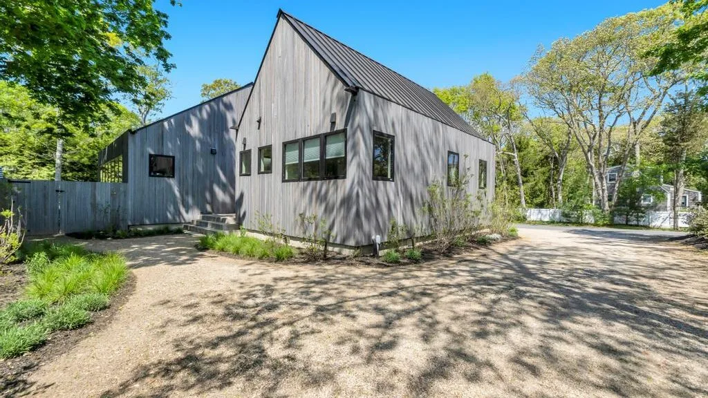 Una casa gris moderna se encuentra junto a un camino de grava, rodeada de árboles.