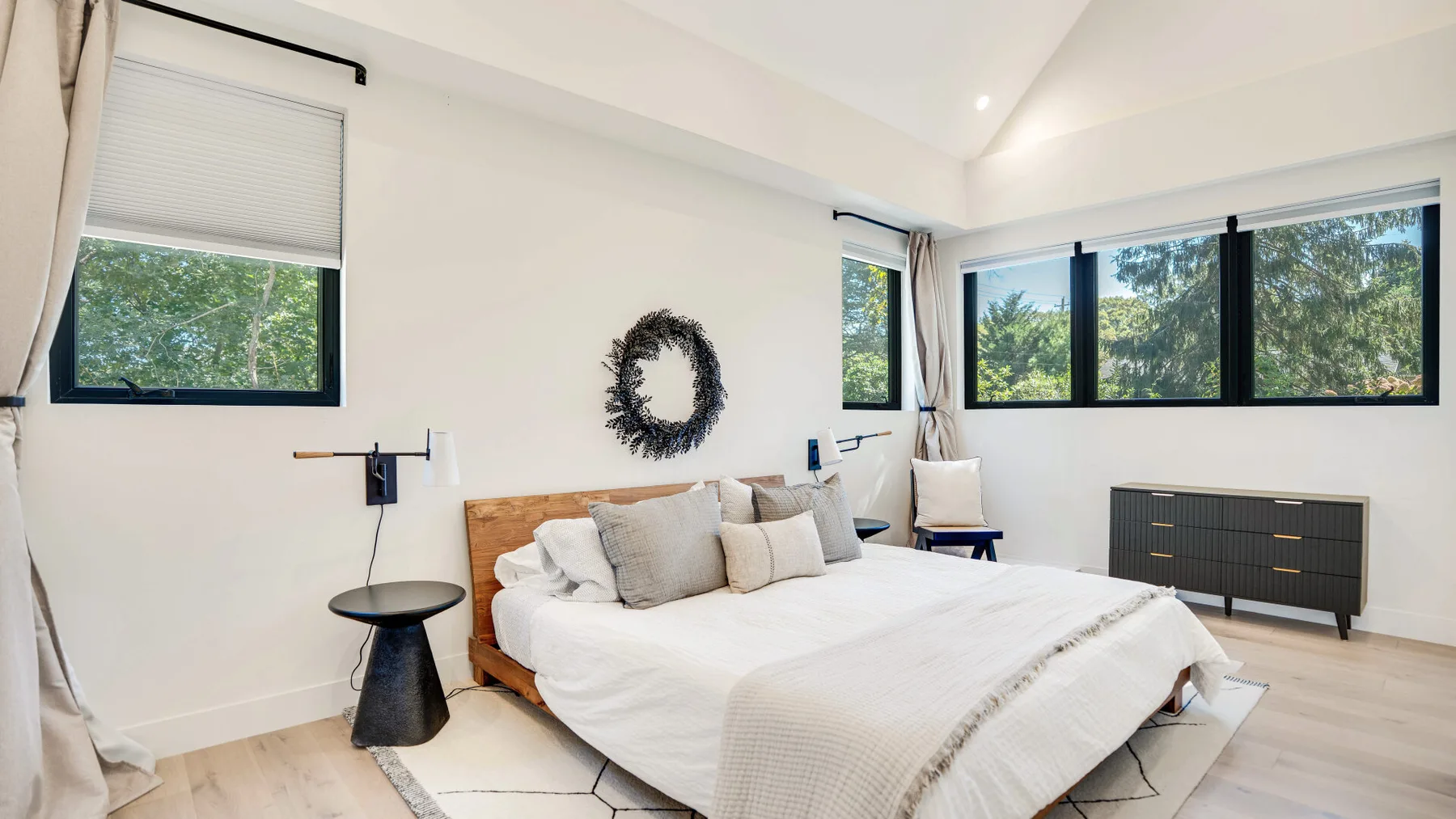 Un dormitorio luminoso con una cama baja, ropa de cama clara y ventanas grandes.
