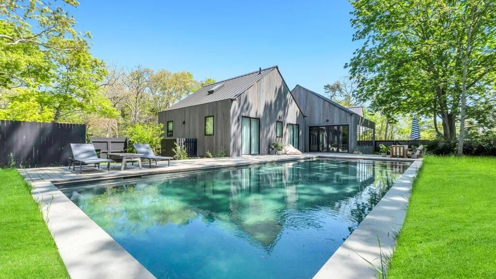 Una casa moderna está junto a una piscina rectangular rodeada de árboles.