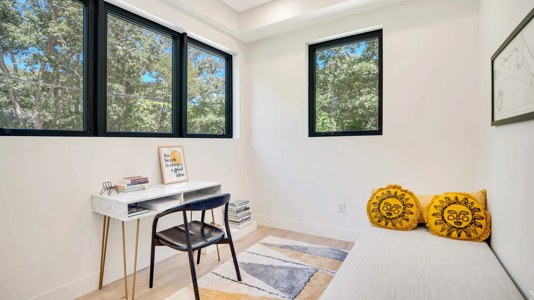 Un dormitorio luminoso tiene una cama con cojines amarillos, un escritorio y ventanas grandes.