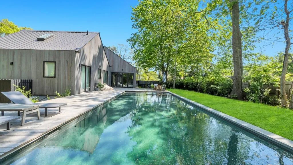 Una casa moderna y un patio bordean una piscina rodeada de árboles y césped.