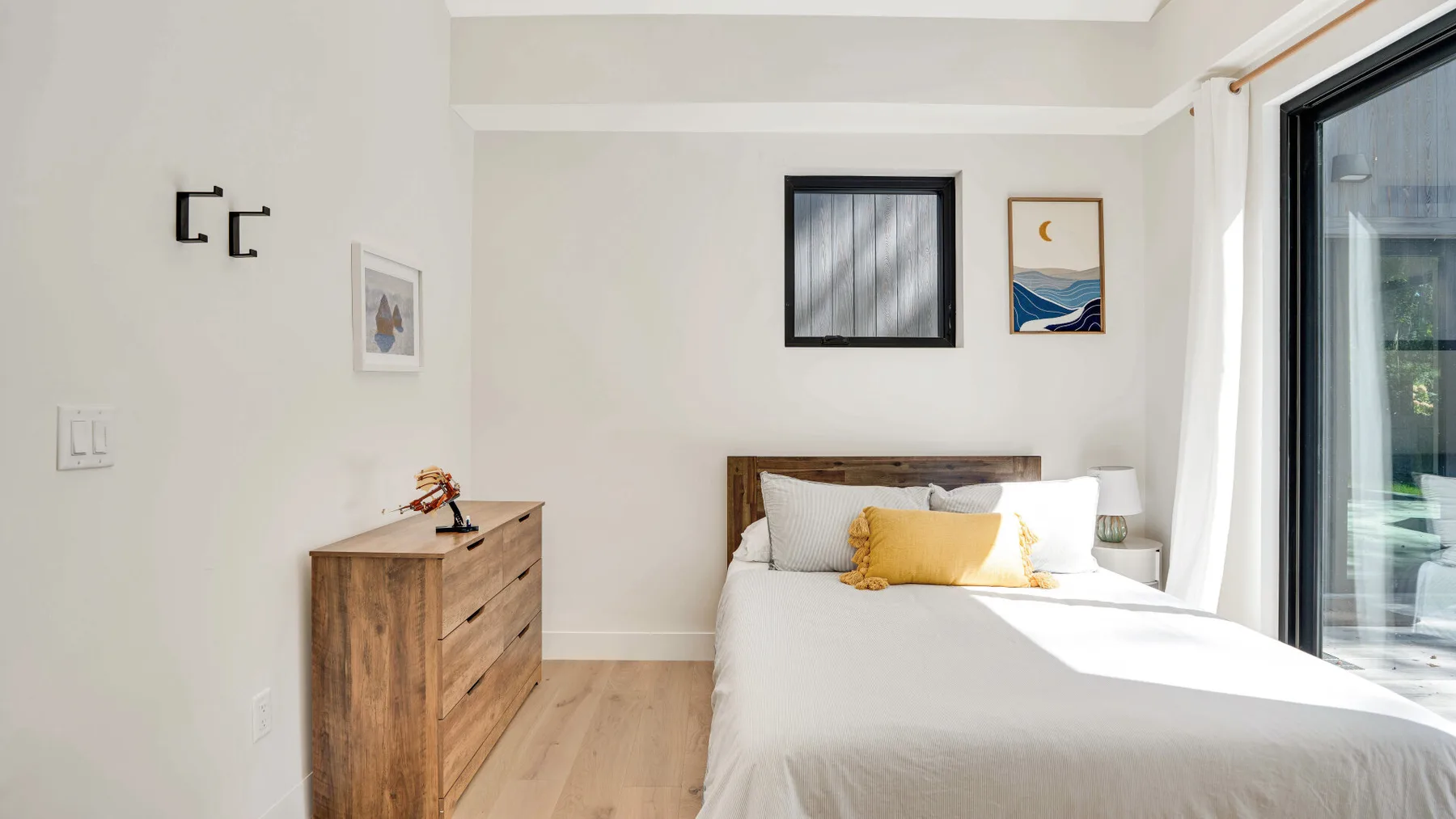 Un dormitorio luminoso con una cama, una cómoda de madera y grandes puertas de vidrio.