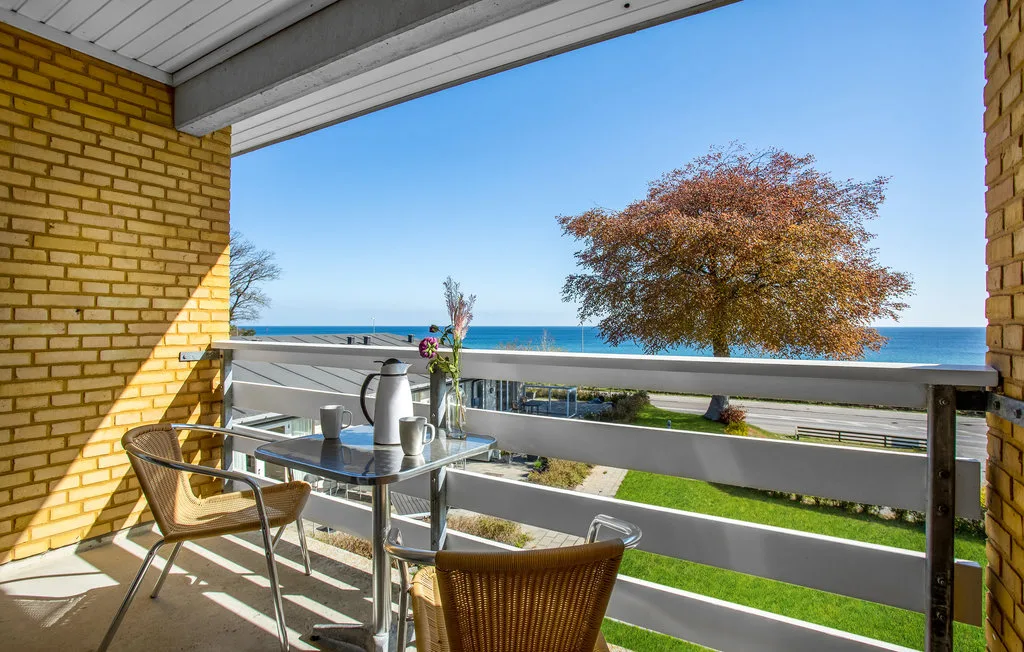 A balcony with outdoor seating overlooks a tree, roadway, and the sea.