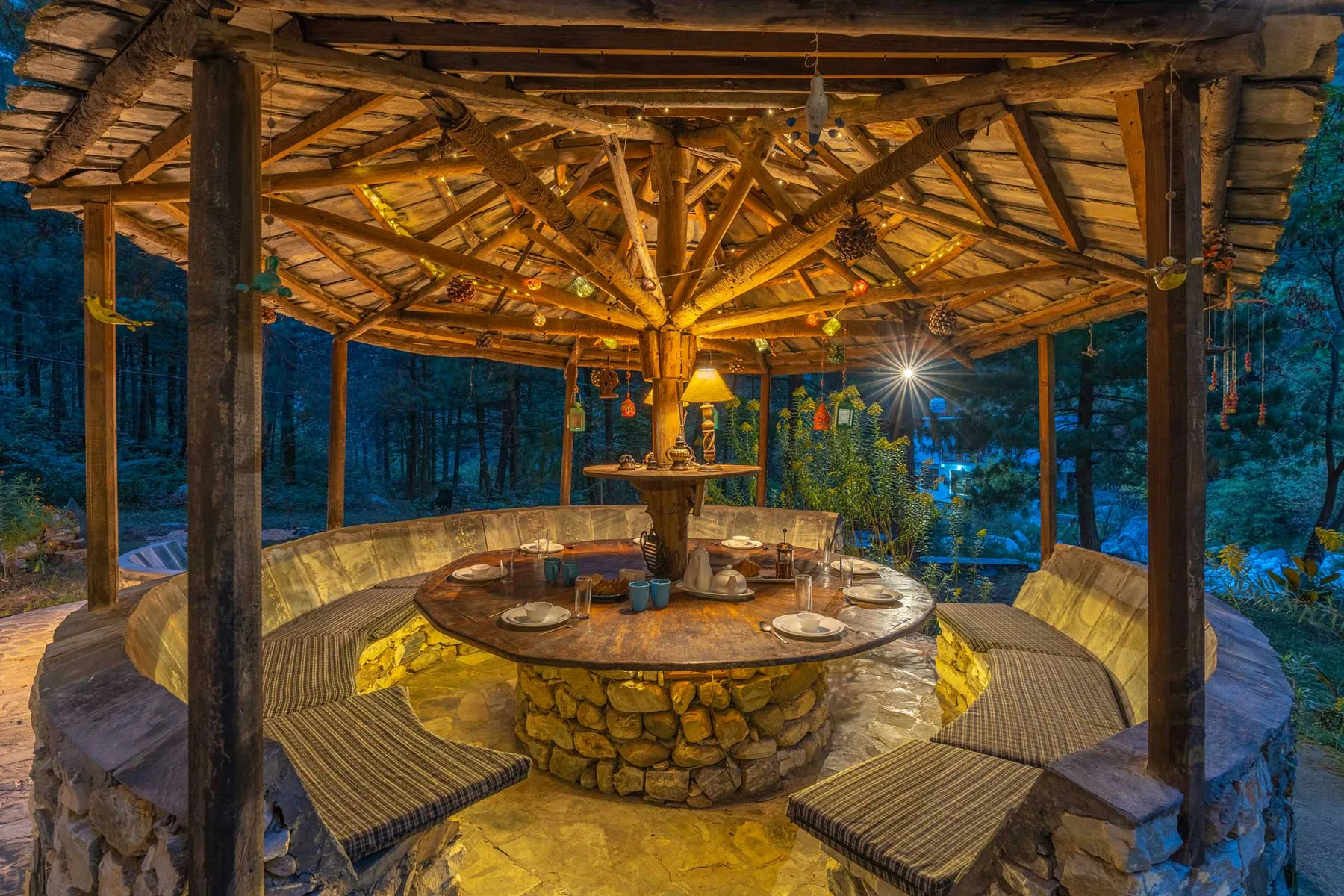 Una zona de estar cubierta al aire libre rodea una mesa redonda de piedra bajo un techo de madera.