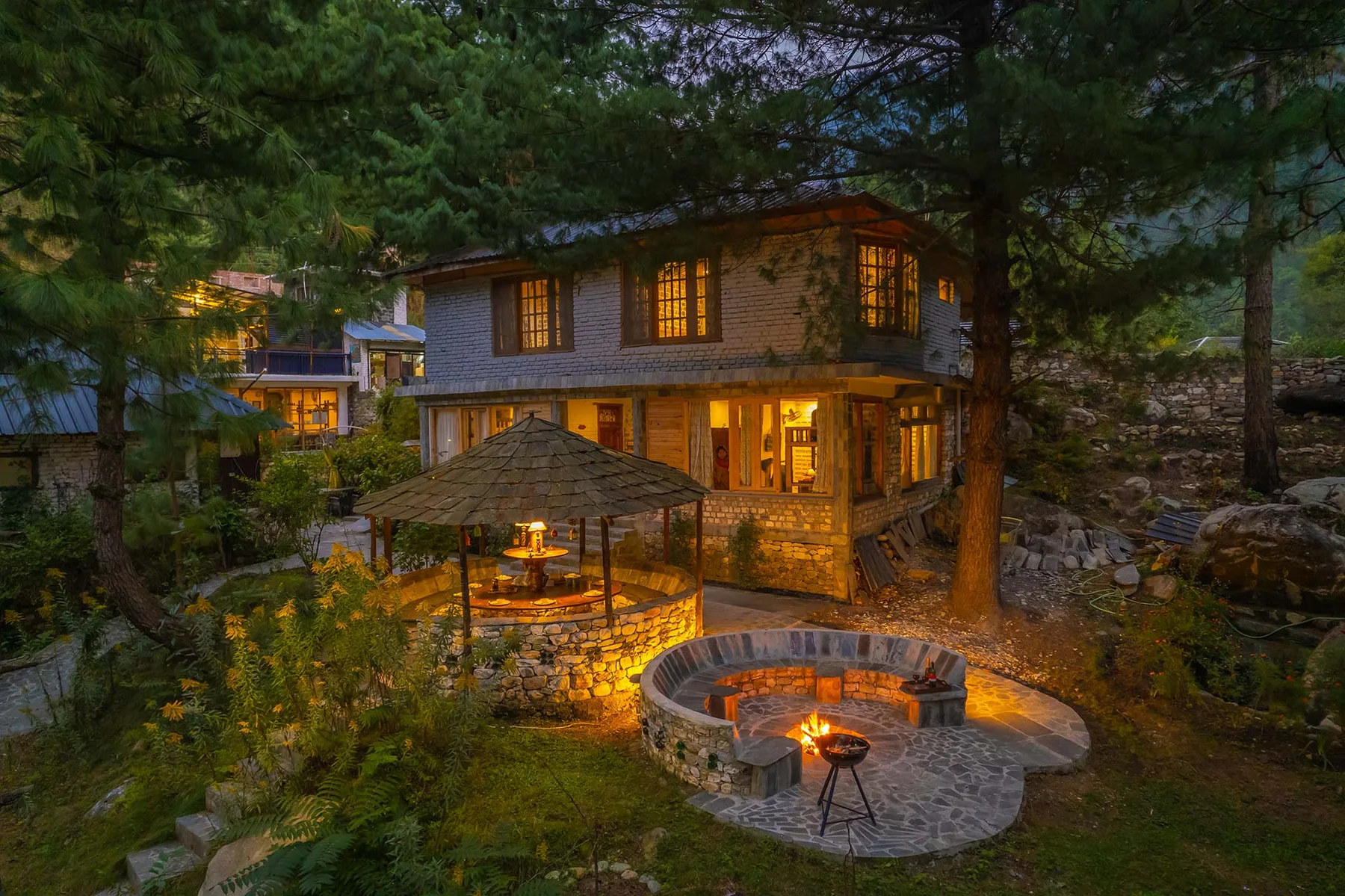 Una casa iluminada se alza entre árboles junto a una zona circular de piedra con una fogata.