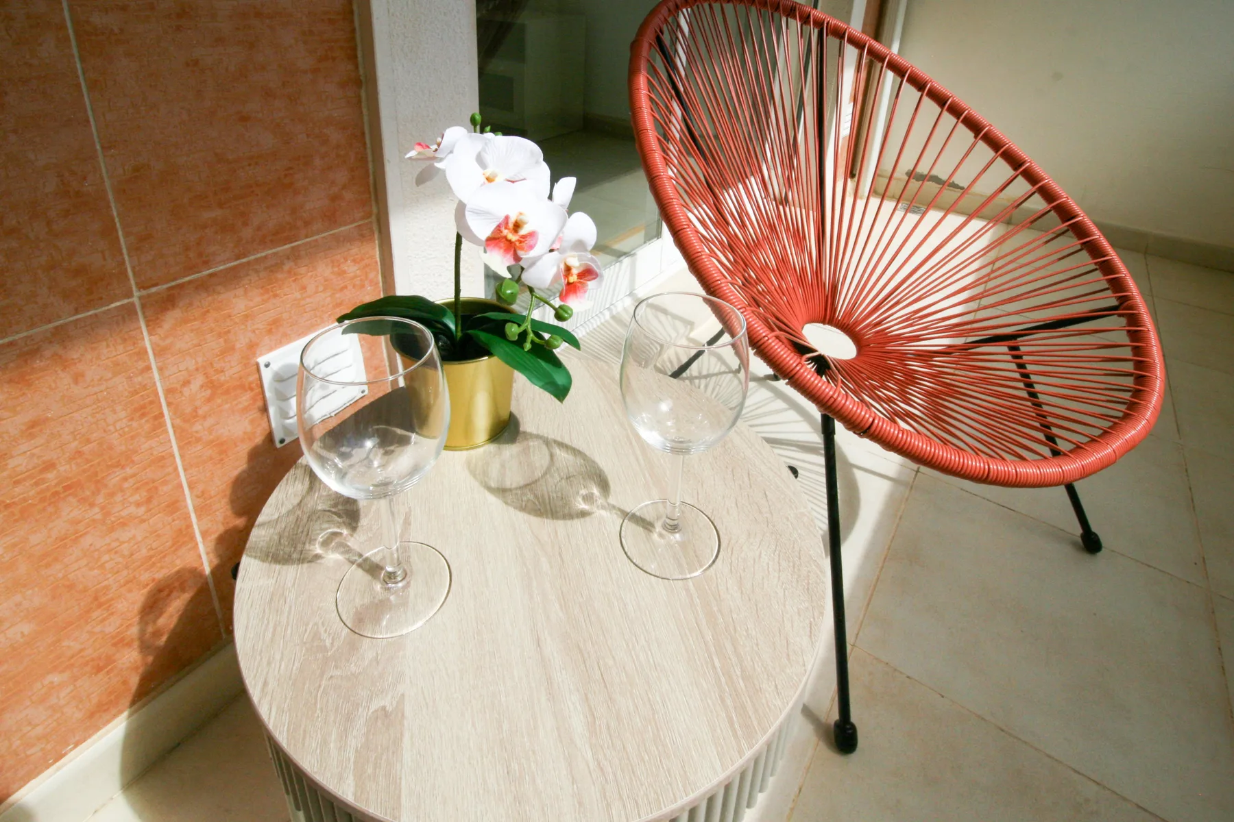 Una mesa pequeña con flores y copas está junto a una silla roja trenzada.